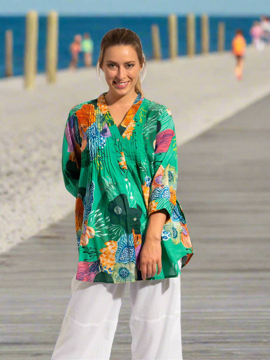 A woman stands on a wooden boardwalk at the beach wearing a vibrant green short-sleeve button-up top with a colorful tropical floral print and white pants.