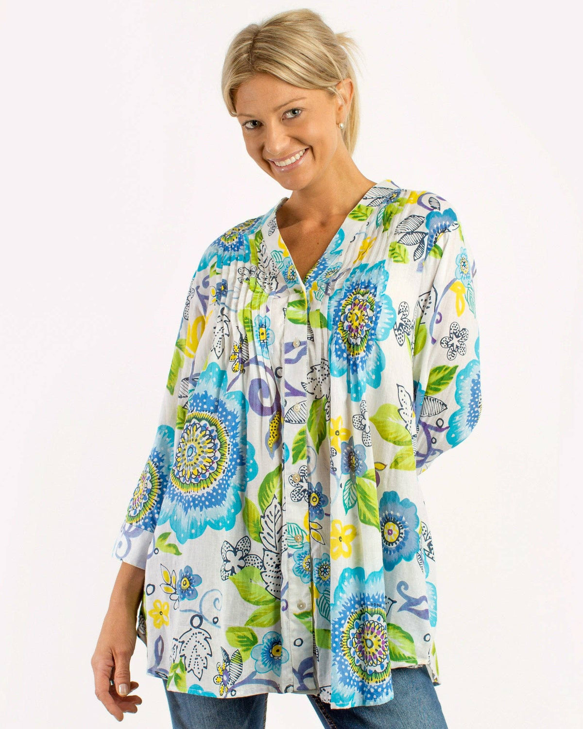 A woman wearing a white long-sleeve tunic top with a colorful floral and mandala print in blue, green, yellow, and purple, paired with blue jeans, against a plain white background.