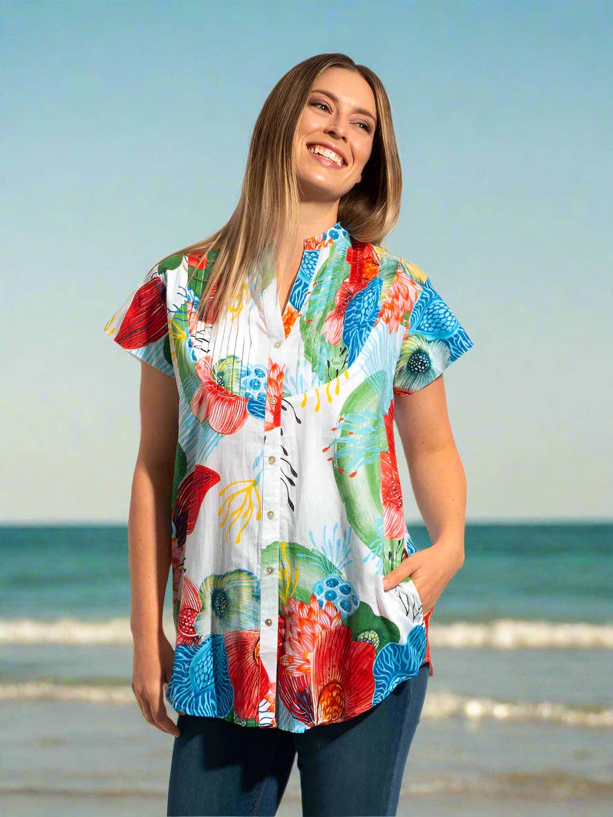 A woman wearing a colorful short-sleeve button-up shirt with a vibrant abstract print, standing on a beach with the ocean and sky in the background.