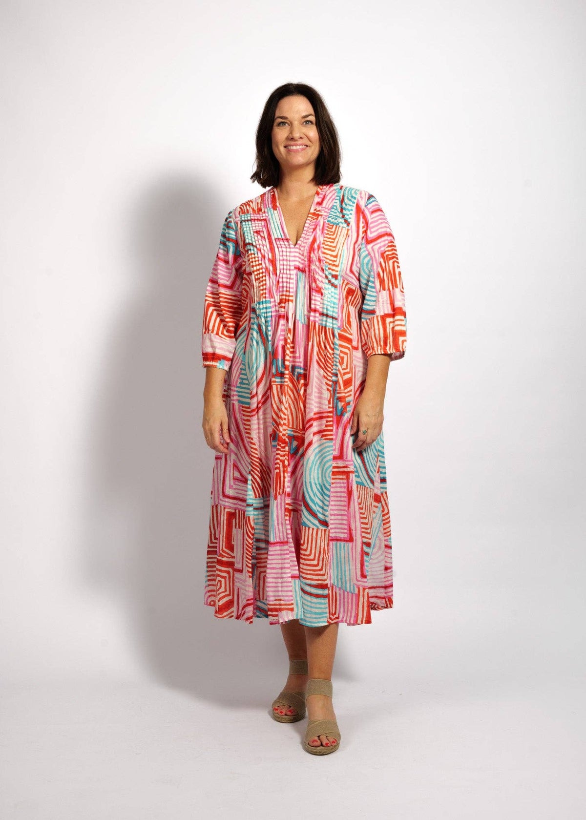 A woman modeling a long-sleeve midi dress with a vibrant abstract geometric pattern in coral, orange, pink, and teal, standing against a light gray studio background.