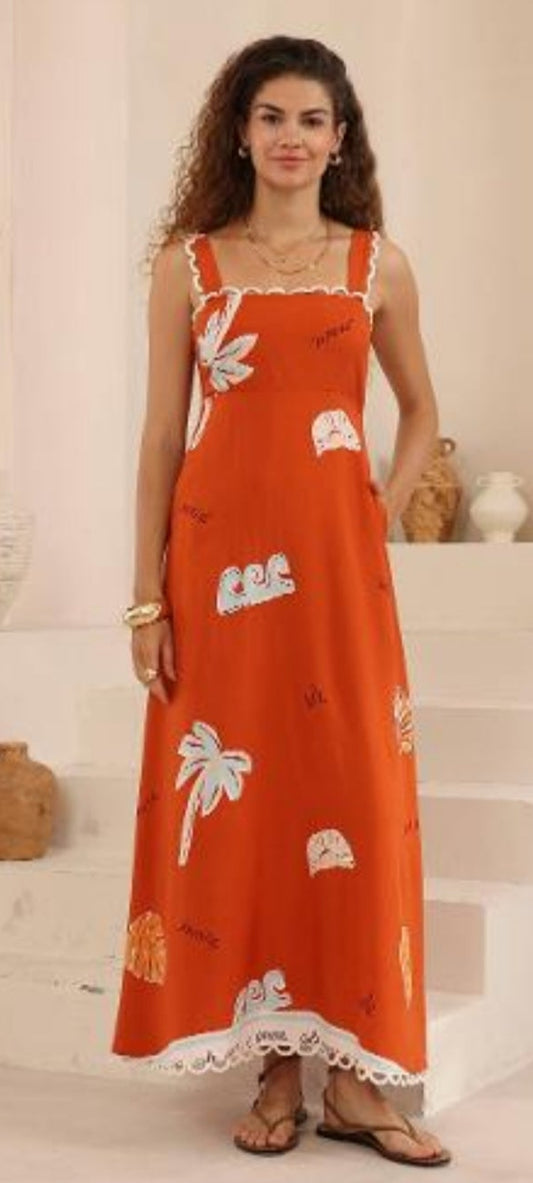 A woman modeling a sleeveless orange maxi dress with white palm tree and sun motifs, standing in a bright, beige-toned interior setting.