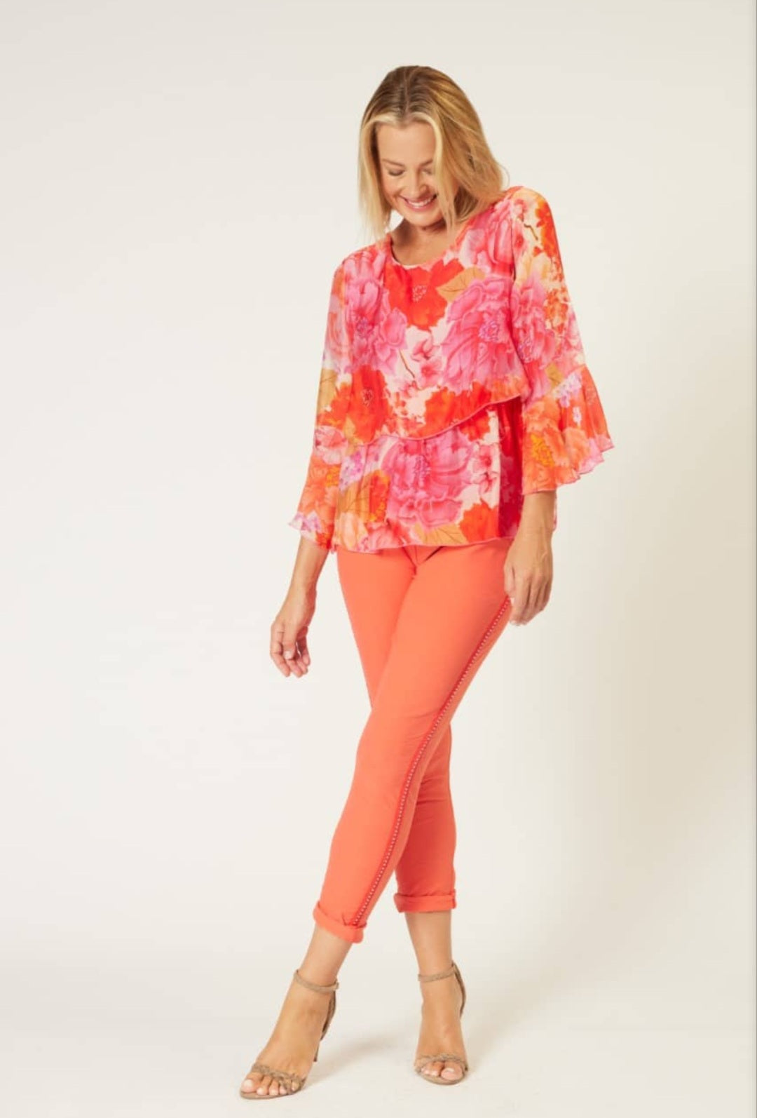 A woman modeling a vibrant floral long-sleeve top in orange, pink, and red, paired with bright orange leggings and beige high-heeled sandals, against a plain light background.