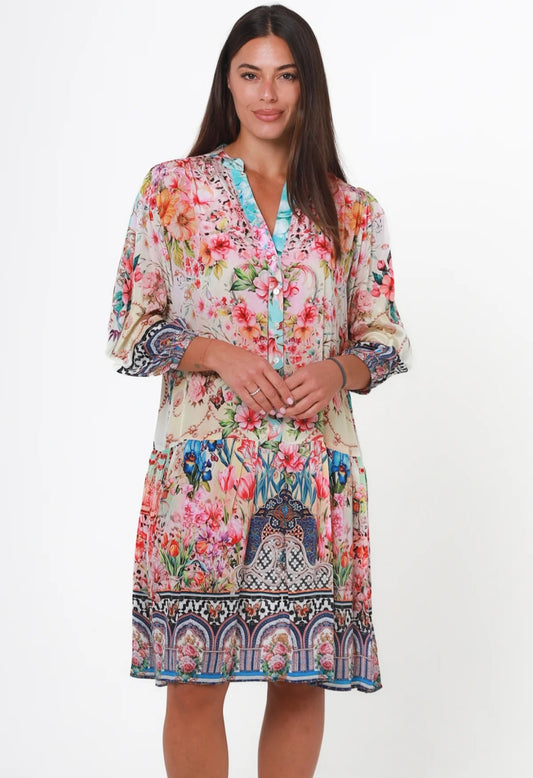 A woman modeling a knee-length bohemian floral print dress with 3/4 sleeves, a V-neckline, and a decorative border print at the hem, set against a plain white studio background.