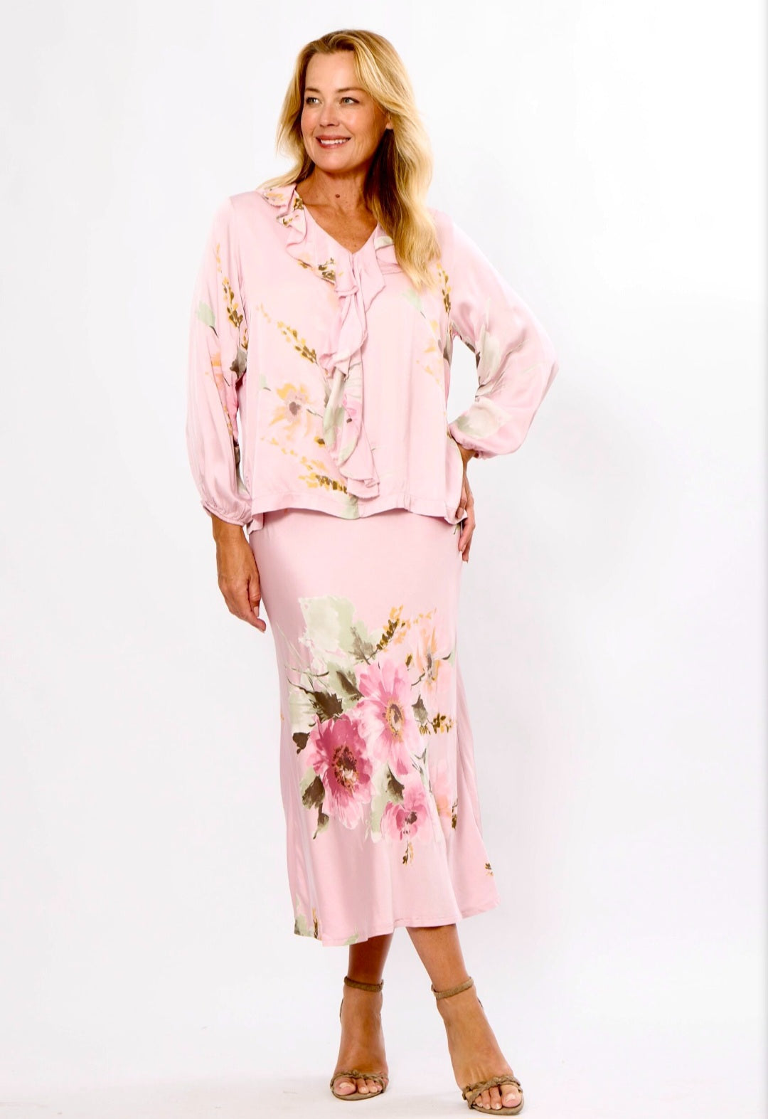 A woman modeling a light pink two-piece set featuring a long-sleeve blouse and matching midi skirt, both decorated with a pink and green floral print, set against a plain white studio background.