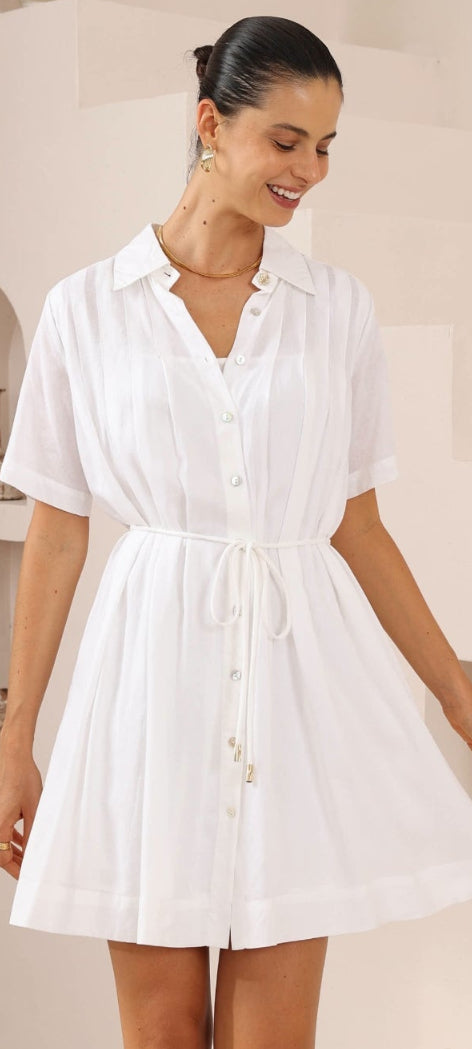 A woman wearing a white short-sleeve collared shirt dress with a tie waist, shown in an indoor setting with cream-colored walls and shelves. The image appears to be a screenshot from a retail listing page with product code text and promotional text at the bottom.