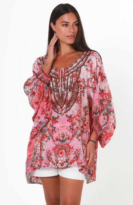 A woman modeling a loose, long-sleeve bohemian-style tunic top with a pink, red, and green floral print, paired with white shorts, against a plain white background.
