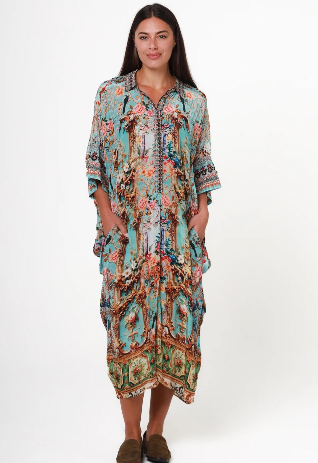 A woman modeling a long, loose kaftan dress with a colorful bohemian floral and paisley print on a light turquoise background, set against a plain white studio background.