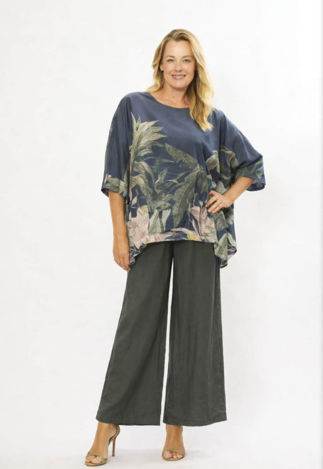 A woman modeling a loose-fitting short-sleeve top with a blue tropical palm leaf print and dark charcoal wide-leg pants, standing against a plain white studio background.