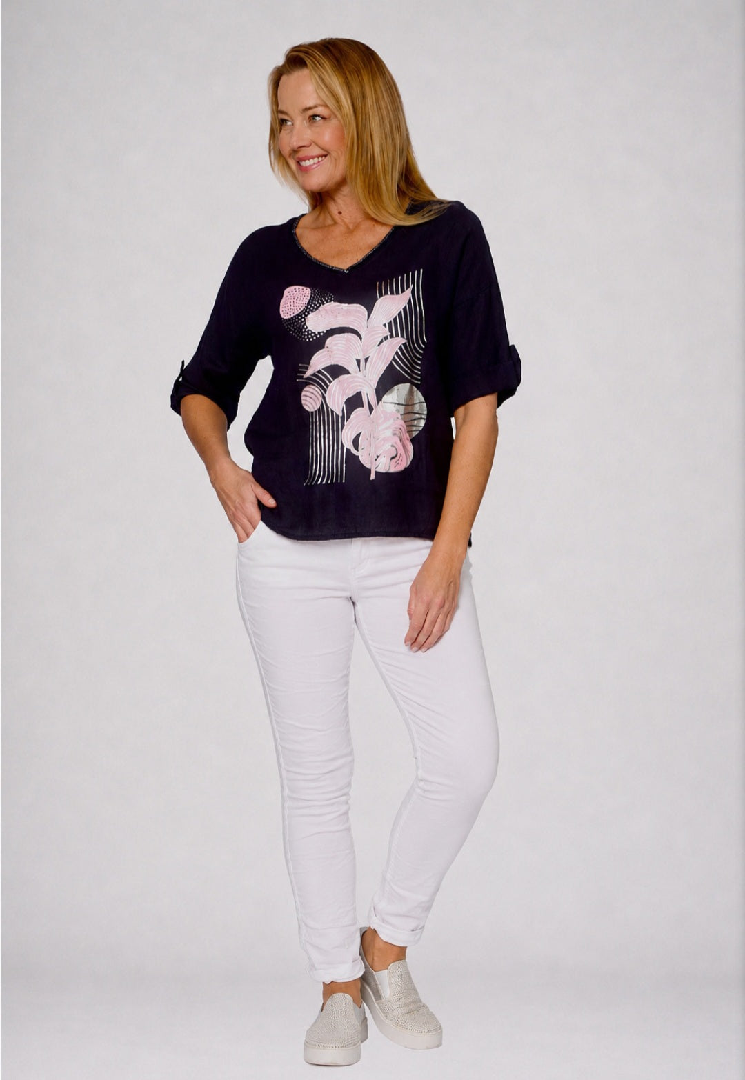 A woman modeling a dark navy blue top with a pink and white graphic print, paired with white pants and light gray sneakers, against a light gray studio background.
