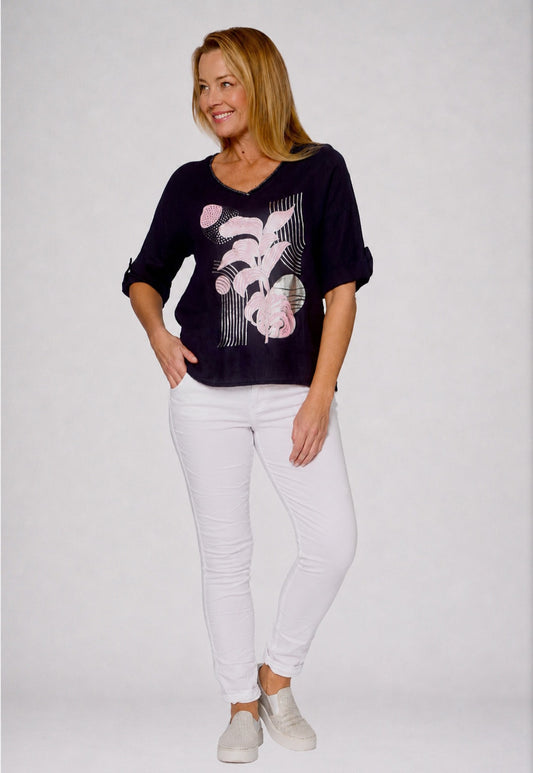 A woman modeling a dark navy blue top with a pink and white graphic print, paired with white pants and light gray sneakers, against a light gray studio background.