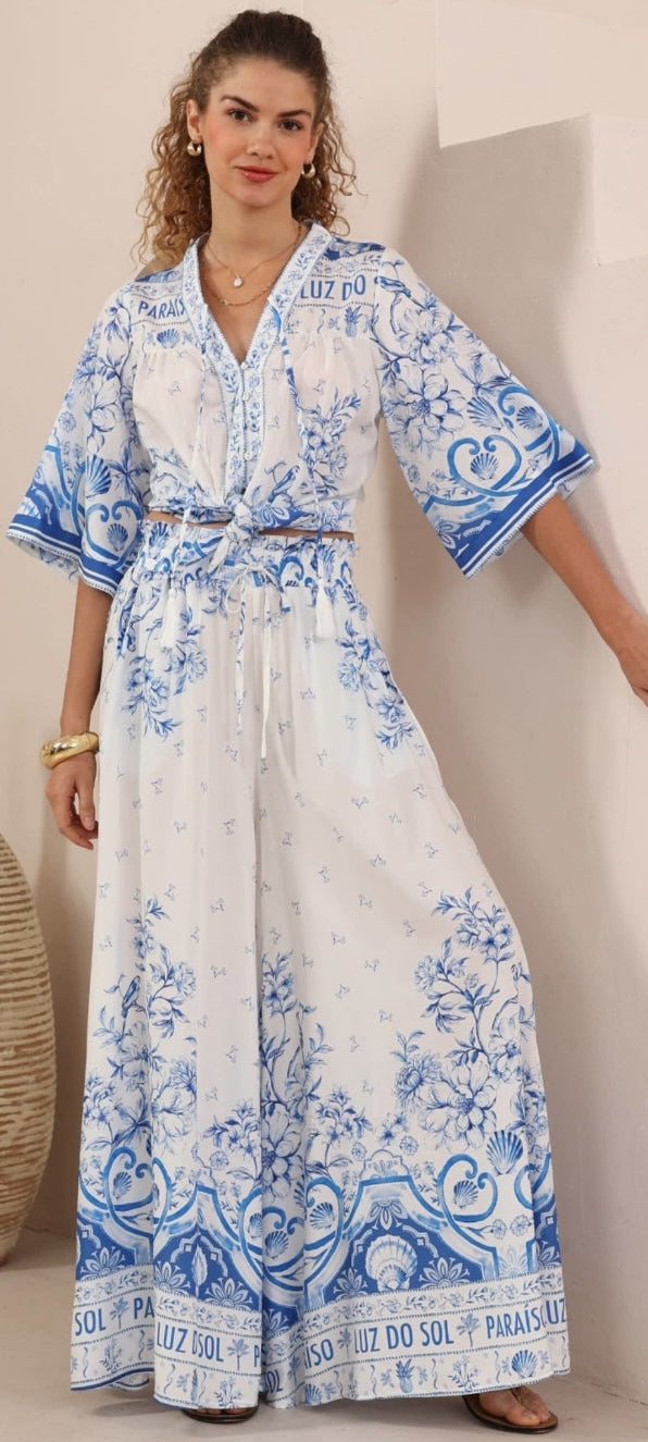 A woman modeling a white maxi kaftan dress with blue floral and paisley patterns, standing in a brightly lit room with a beige vase in the background.
