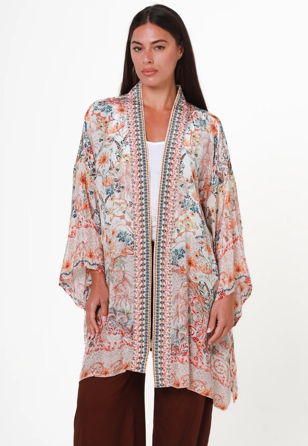 A woman modeling a lightweight open-front kimono-style cardigan with a beige base and a multicolor floral and paisley print in orange, blue, and green, styled over a white top and brown pants against a plain white background.